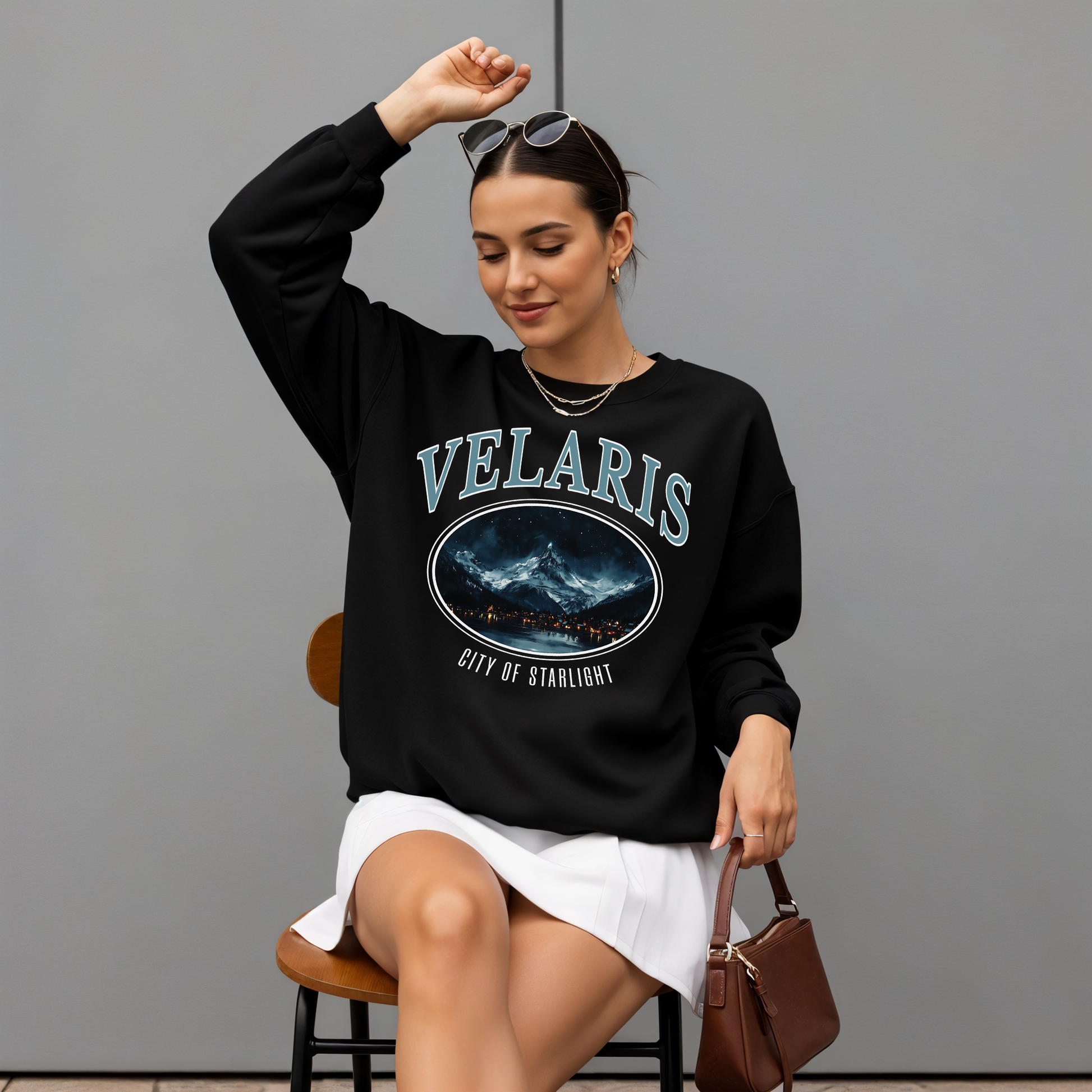 Woman wearing a black 'VELARIS' sweatshirt with a graphic design, sitting on a chair against a gray background.