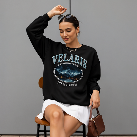 Woman wearing a black 'VELARIS' sweatshirt with a graphic design, sitting on a chair against a gray background.